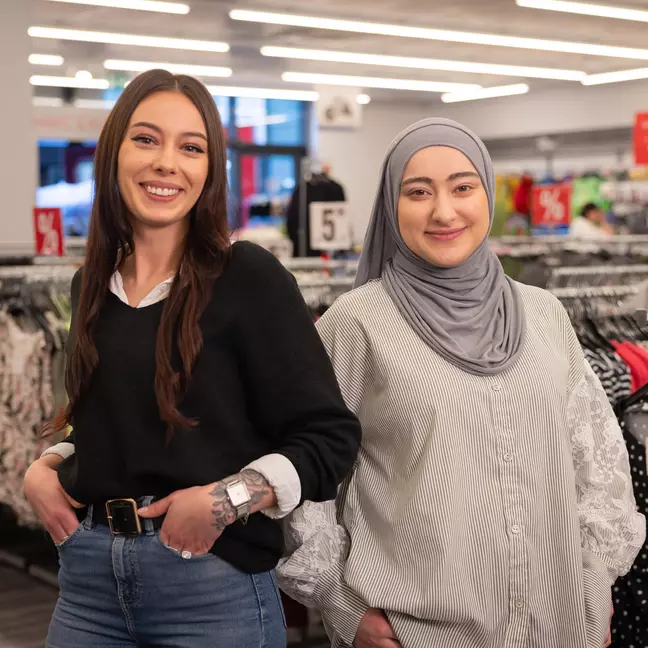Két munkatársunk mosolyog a ruhaállványok előtt egyik KiK-üzletünkben