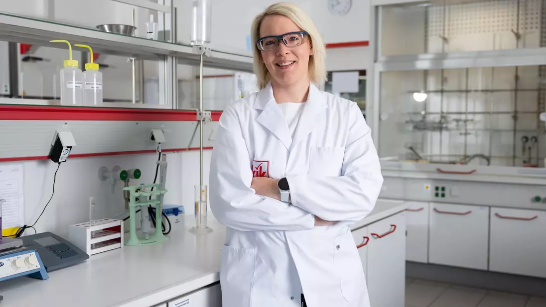 Egy fehér köpenyes laboráns karba tett kézzel áll egy modern laboratóriumban, védőszemüveget visel és mosolyog.
