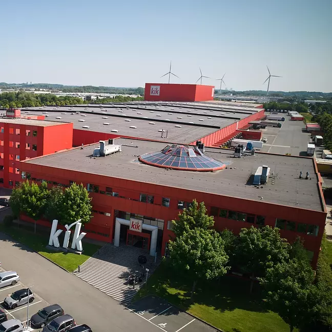 Egy nagy, piros cégépület, amelyen a „kik” logó látható, egy zöld táj közepén áll.