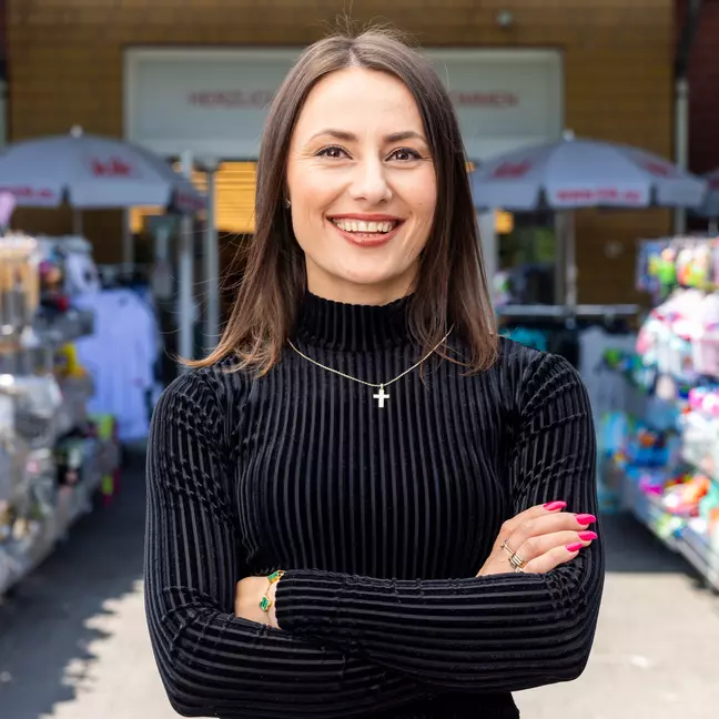 Egy munkatárs az üzlet előtti kültéri részen áll az áruhordozók között, és mosolyogva néz a kamerába.