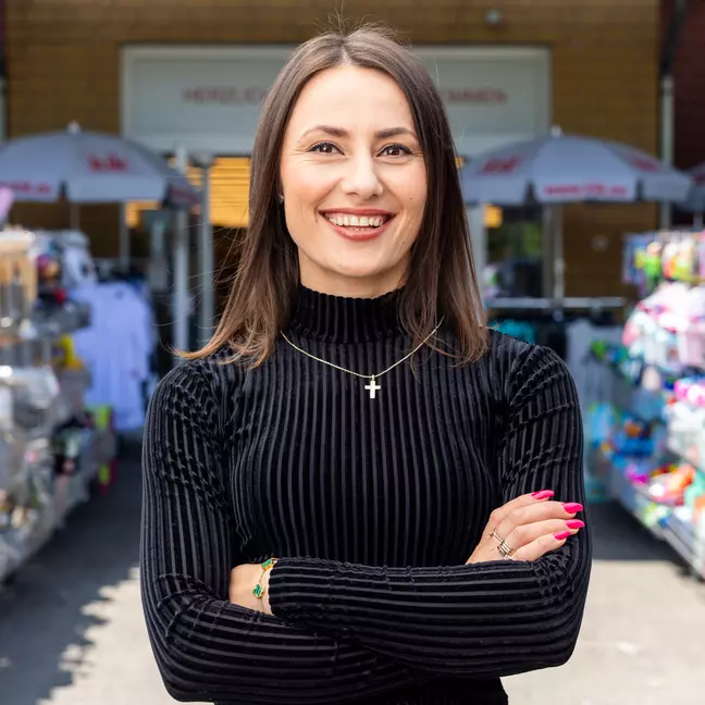 Egy munkatárs az üzlet előtti kültéri területen áll az áruhordozók között, és mosolyogva néz a kamerába.